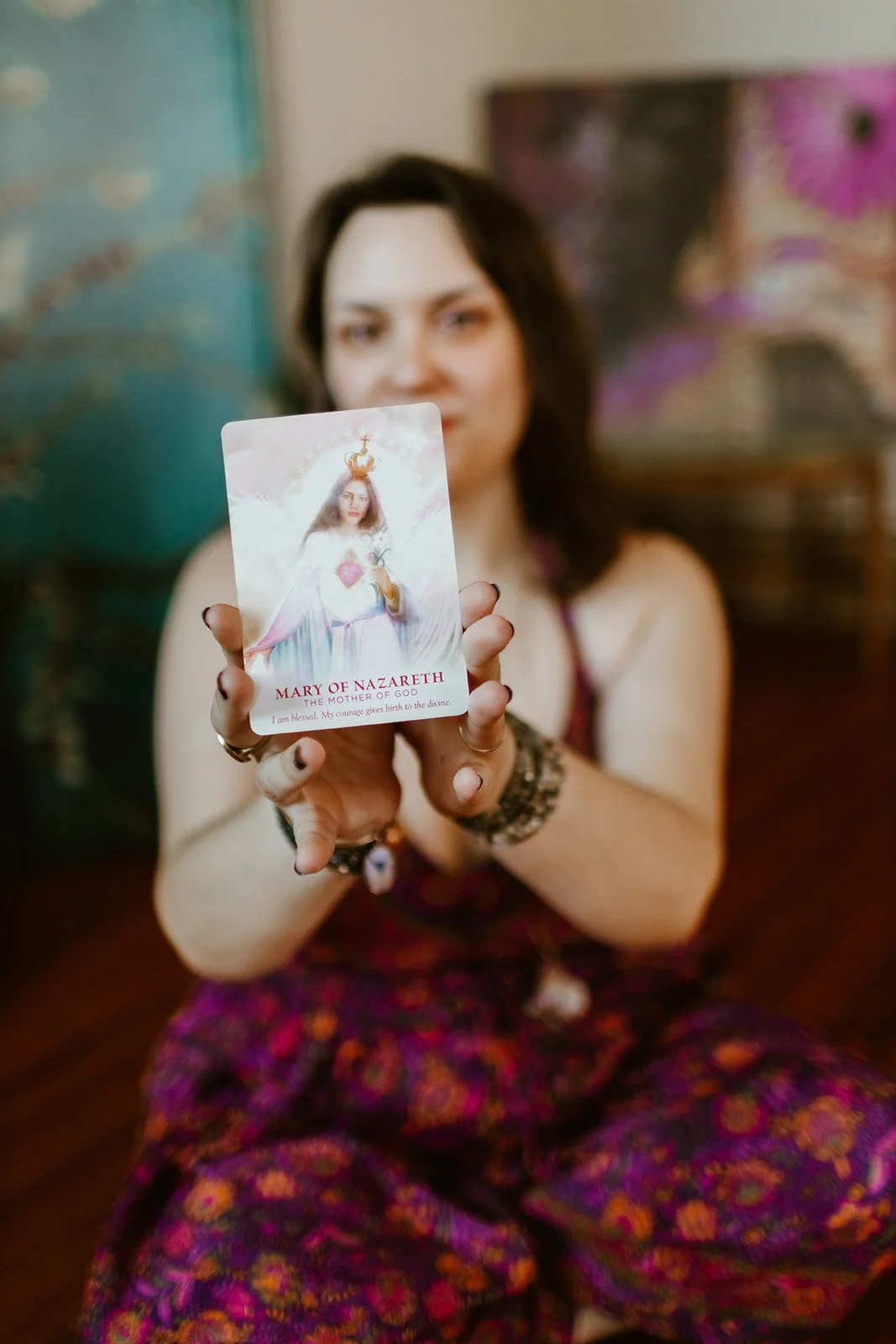 A card of Mary of Nazareth, Mother of God, is held up in focus in the foreground. Author Safrianna Lughna holds the card, off focus in the background, wearing a floral jumper. Photograph by Amanda Lucia Photography.