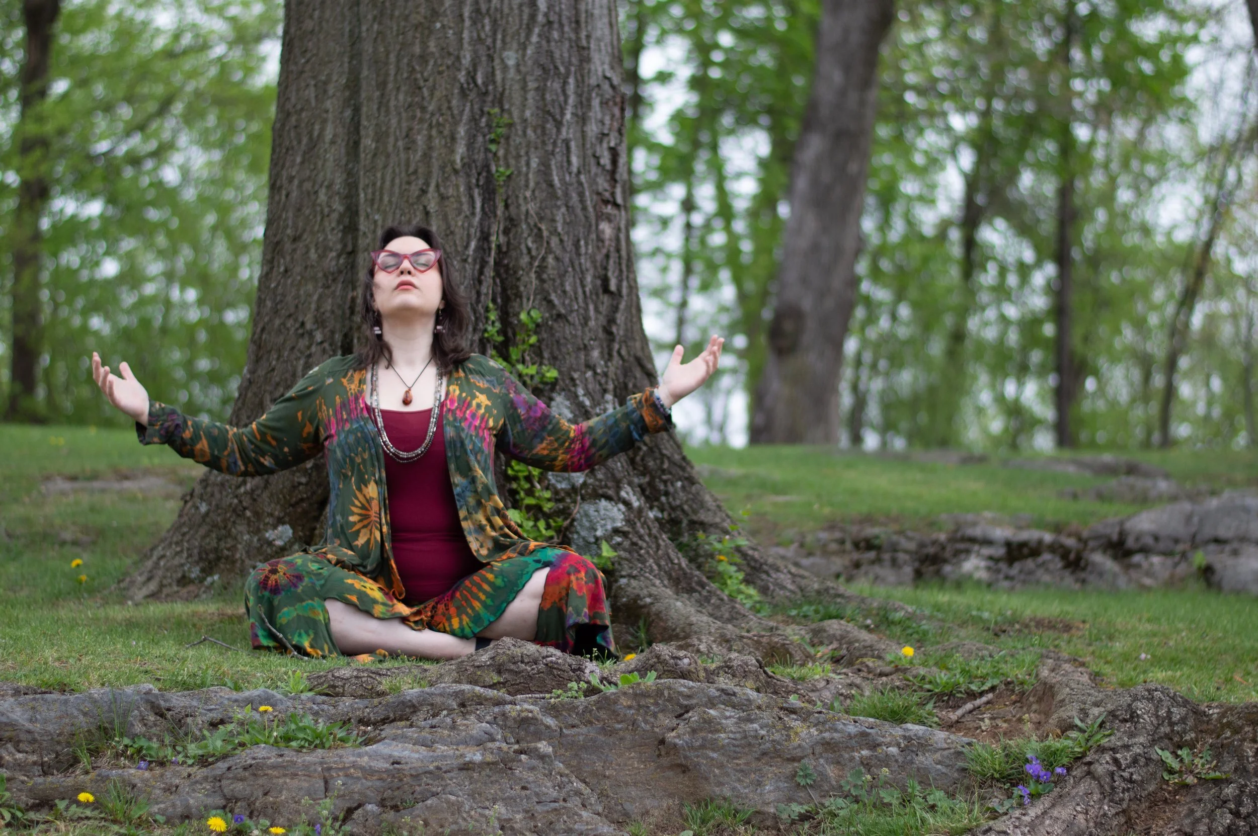 Safrianna sits before a large tree with her arms uplifted in meditation